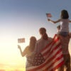 Parents and two daughters, draped in a flag, with the girls waving little flags, look at a beautiful sunset.