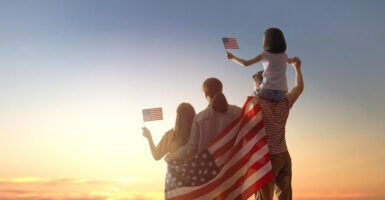 Parents and two daughters, draped in a flag, with the girls waving little flags, look at a beautiful sunset.