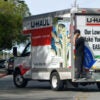 A man in black chats on his cellphone while standing on the back ledge of a Uhaul truck that's pulling out on a suburban street.