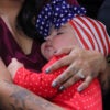 A dark haired mother with tattooed arms holds an infant dressed in red-and-white with its head wrapped in an American flag cap.