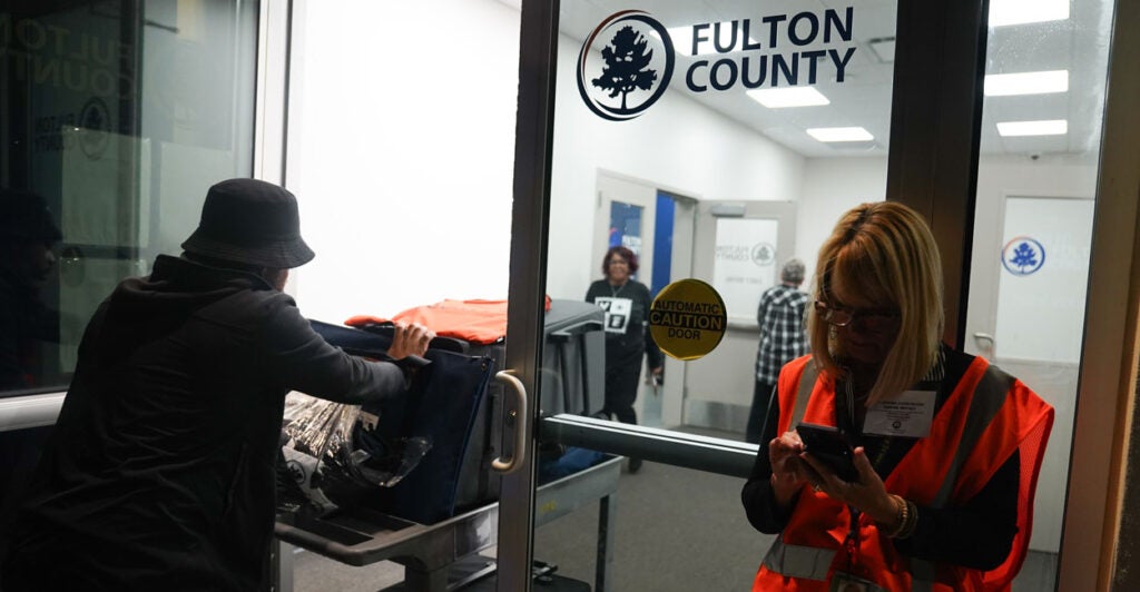An election worker pushing a cart of ballots goes through a glass door that reads "Fulton County."