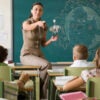 A female teacher sits on a desk in front of a blackboard pointing toward her students.