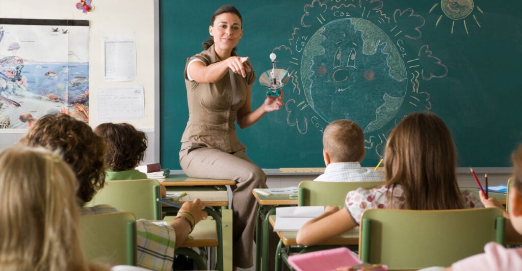 A female teacher sits on a desk in front of a blackboard pointing toward her students.