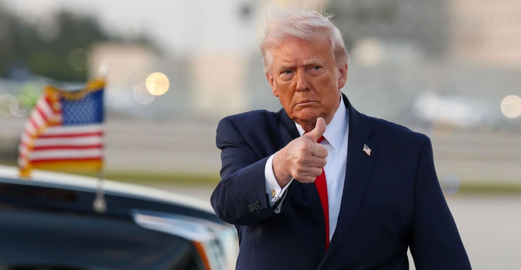 President Donald Trump offers a thumb's up, with his limousine parked behind him.