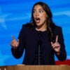 Alexandria Ocasio-Cortez in black suit with eyes and mouth wide, both hands pointing upwards speaks at podium at DNC convention.