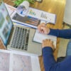 Woman in blue shirt takes notes on a notepad in front of laptop computer with graphs on paper on both sides.