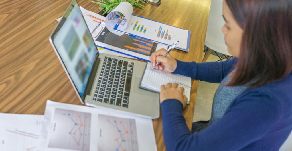 Woman in blue shirt takes notes on a notepad in front of laptop computer with graphs on paper on both sides.