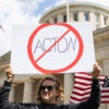 A woman stands outside of the Ohio capitol with a prohibition slash through symbol sign of "Acton"