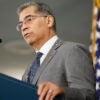 Health and Human Services Secretary Xavier Becerra stands at a podium with an American flag in the background.