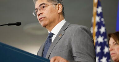 Health and Human Services Secretary Xavier Becerra stands at a podium with an American flag in the background.