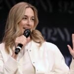 A woman with long blonde hair, wearing a white jacket, speaks into a microphone and gestures with her left hand on stage. The background features blurry 92NY text.